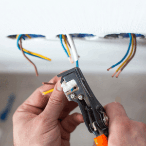 Mains d'électricien dénudant des fils électriques (marron, jaune) avec une pince, câbles sortant d'un plafond blanc.