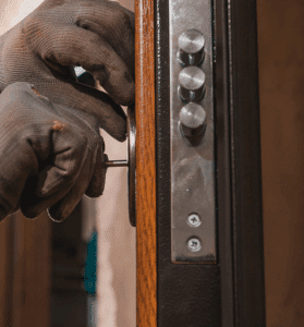 Mains gantées manipulant une clé dans une serrure de porte de sécurité en bois et métal, avec trois verrous apparents.