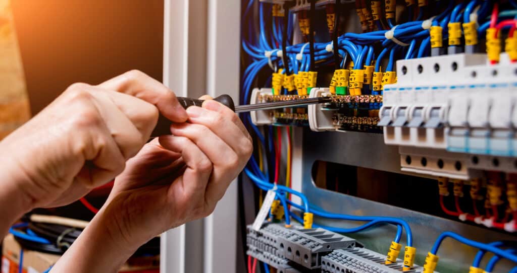Mains d'électricien ajustant un fil dans un tableau électrique. Câblage bleu, jaune numéroté, disjoncteurs.