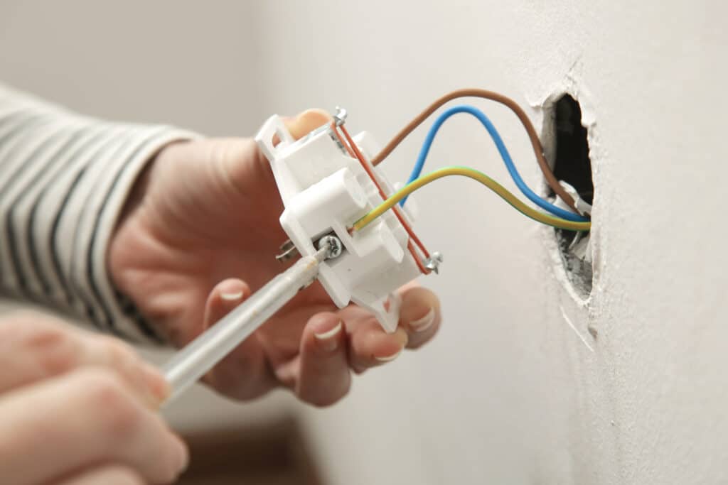 Mains installant une prise électrique murale blanche. Un tournevis connecte les fils colorés (marron, bleu, jaune-vert) sortant du mur.