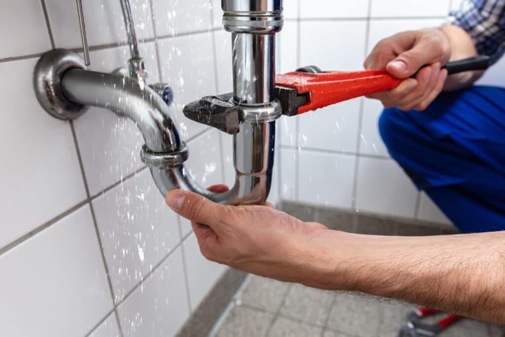 Plombier répare une fuite d'eau sur un tuyau d'évier chromé avec une clé à molette rouge. L'eau gicle sur le mur carrelé.